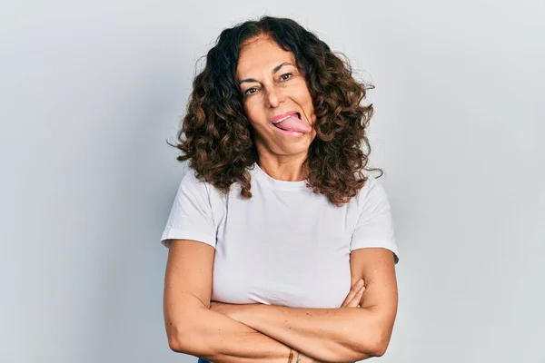 Middle age hispanic woman with arms crossed gesture sticking tongue out happy with funny expression.