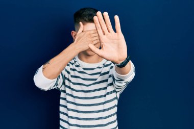 Kulakları genişlemiş, sıradan çizgili tişört giyen, gözlerini ellerle kapayan, üzgün ve korku ifadeli hareketler yapan beyaz bir çocuk. utangaç ve olumsuz kavram. 