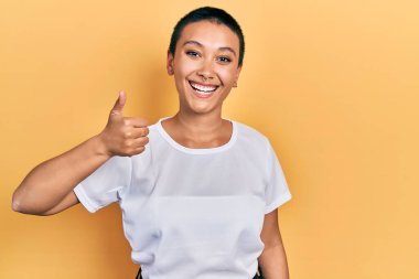 Kısa saçlı, sıradan beyaz tişörtlü güzel İspanyol kadın mutlu ve pozitif gülümsüyor, başparmağıyla mükemmel ve onay işareti yapıyor. 
