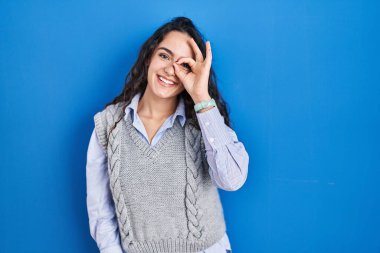 Genç esmer kadın mavi arka planda duruyor. El hareketleriyle gülümsüyor, gözleriyle mutlu yüzüyle parmaklarının arasından bakıyor.. 