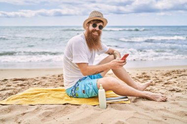 Genç kızıl saçlı adam kumsalda havluya oturmuş akıllı telefon kullanıyor..