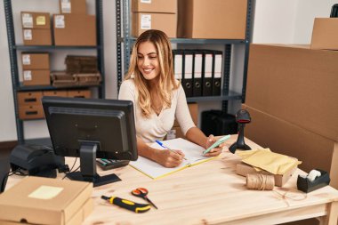 Genç sarışın kadın ecommerce iş işçisi akıllı telefon kullanıyor ve ofiste kitap yazıyor.