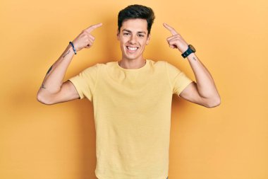 Gündelik sarı tişört giyen genç İspanyol adam iki eliyle başını işaret ederek gülümsüyor, harika bir fikir ya da düşünce, iyi hafıza 