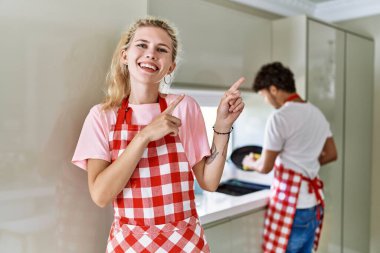 Young caucasian woman wearing apron and husband doing housework washing dishes smiling and looking at the camera pointing with two hands and fingers to the side. 