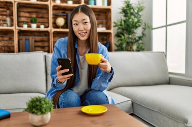 Genç Çinli kız akıllı telefon kullanıyor ve evdeki kanepede oturup kahve içiyor..