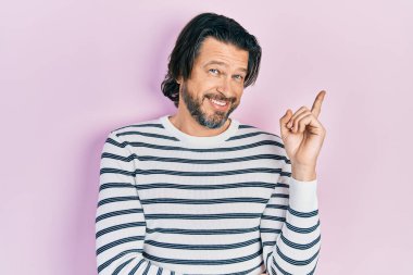 Orta yaşlı, günlük kıyafetler giyen, yüzünde kocaman bir gülümseme olan beyaz bir adam, kameraya el ve parmak ile bakıyor.. 