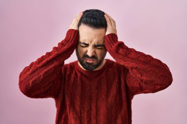 Sakallı, pembe arka plan üzerine kazak giyen, çaresiz ve stresli, acı ve migrenden muzdarip genç İspanyol bir adam. Eller başının üstünde.. 