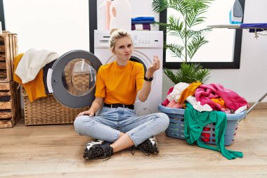 Young blonde woman doing laundry sitting by washing machine doing italian gesture with hand and fingers confident expression 