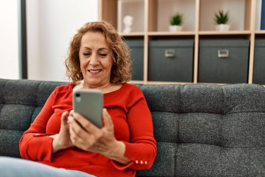 Orta yaşlı beyaz kadın akıllı telefon kullanıyor. Evdeki kanepede oturuyor..