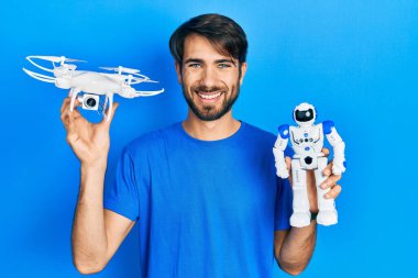 İHA ve robot oyuncağı tutan genç İspanyol adam yüzünde mutlu ve havalı bir gülümsemeyle gülümsüyor. diş gösterme. 
