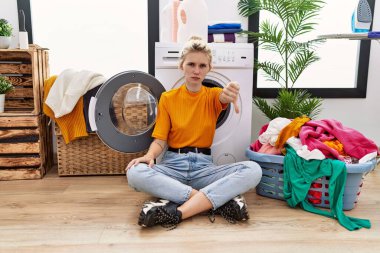 Young blonde woman doing laundry sitting by washing machine looking unhappy and angry showing rejection and negative with thumbs down gesture. bad expression. 