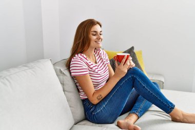 Evdeki kanepede kahve içen genç bir kadın.