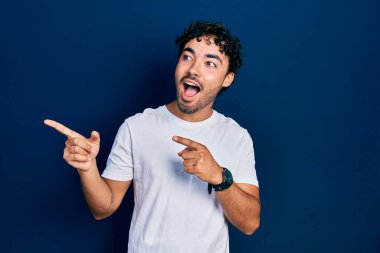 Gündelik beyaz tişört giyen genç İspanyol adam gülümseyip kameraya bakıyor ve iki eliyle ve parmaklarıyla kenarı işaret ediyor.. 