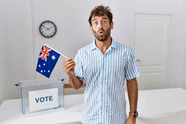 Young handsome man at political campaign election holding australia flag scared and amazed with open mouth for surprise, disbelief face