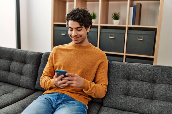 Молодой латиноамериканец с помощью смартфона сидит дома на диване.