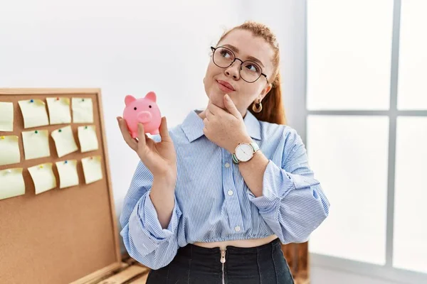 Ung Rödhårig Kvinna Med Spargris Kontoret Allvarligt Ansikte Tänker Fråga — Stockfoto