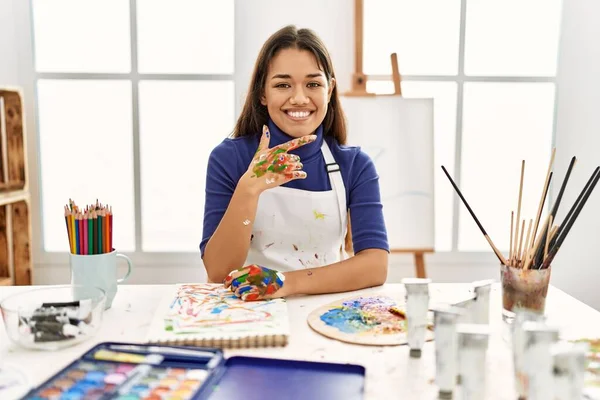 Young brunette woman at art studio with painted hands looking confident at the camera with smile with crossed arms and hand raised on chin. thinking positive.