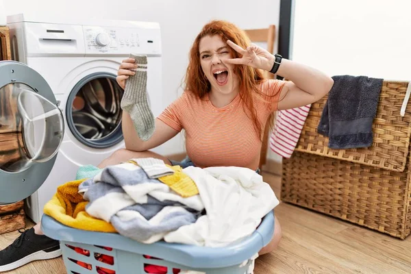 若い赤い頭の女性は顔の上に指で平和のシンボルを行う洗濯機に汚れた洗濯機を入れて 明るい勝利を示す笑みを浮かべて — ストック写真