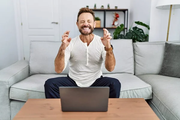 Middle age man using laptop at home gesturing finger crossed smiling with hope and eyes closed. luck and superstitious concept.