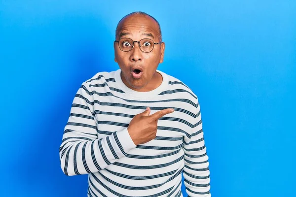 Middle age latin man wearing casual clothes and glasses surprised pointing with finger to the side, open mouth amazed expression.