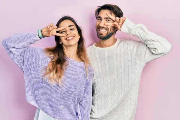 若いヒスパニック系のカップルは 顔の上に指で平和のシンボルを行うカジュアルな服を着て 明るい勝利を示す笑顔 — ストック写真