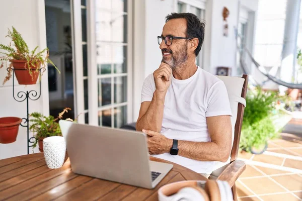 Middle age man using computer laptop at home with hand on chin thinking about question, pensive expression. smiling and thoughtful face. doubt concept.