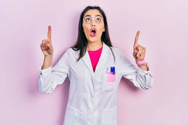Young hispanic girl wearing scientist uniform amazed and surprised looking up and pointing with fingers and raised arms.