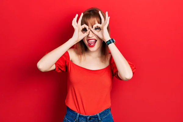 赤毛の若い女性は 双眼鏡の舌を突き出すようなOkジェスチャーを行うカジュアルな赤のTシャツを着て 目の指を見て クレイジーな表現 — ストック写真