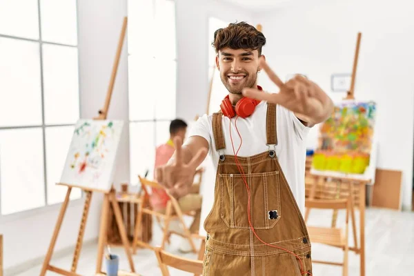 Young hispanic man at art studio smiling looking to the camera showing fingers doing victory sign. number two.