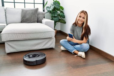 Evde yerde oturan akıllı telefonuyla robot elektrik süpürgesini kontrol eden beyaz bir kız..