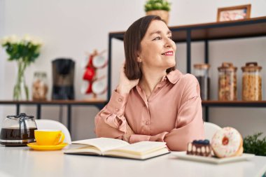 Middle age woman having breakfast reading book sitting on table at home
