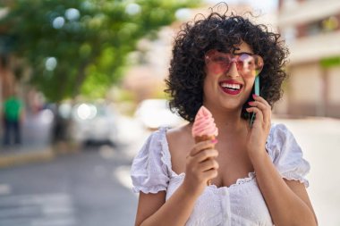 Ortadoğulu genç bir kadın akıllı telefondan konuşuyor. Sokakta dondurma yiyor.