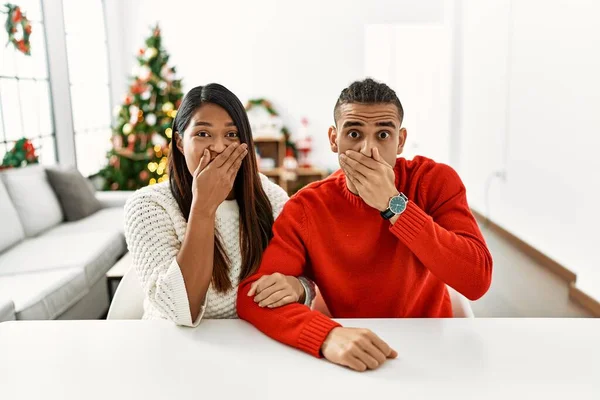 一对年轻的拉丁夫妇坐在圣诞树旁的桌子上 双手捂着嘴 为自己的错误而震惊 秘密概念 — 图库照片