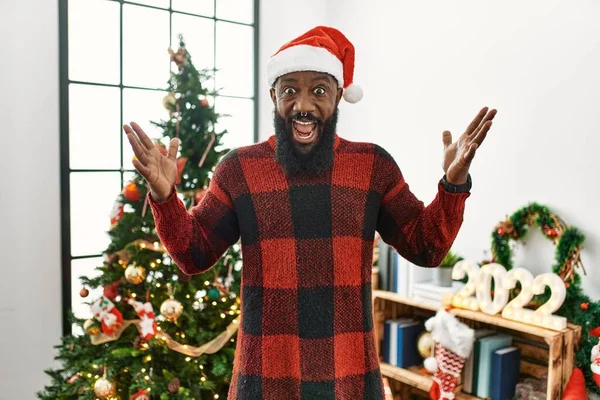 サンタの帽子をかぶったアフリカ系アメリカ人男性がクリスマスツリーのそばに立ってクレイジーを祝い 腕を上げて興奮して叫んで目を開けて成功したことに驚いています 勝者のコンセプト — ストック写真