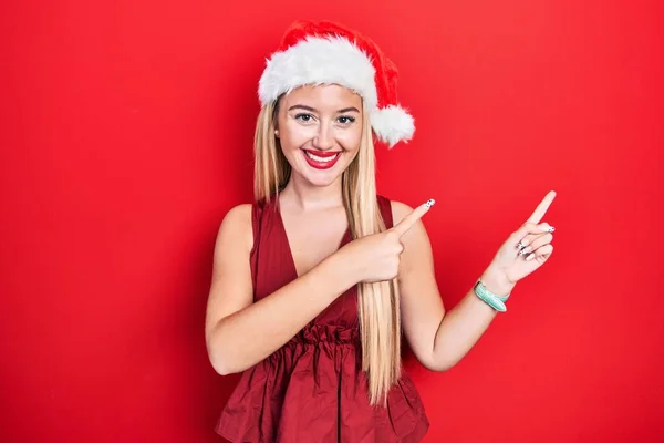 若いですブロンド女の子身に着けていますクリスマス帽子笑顔と見ますザカメラ指しています二つの手と指で側面 — ストック写真
