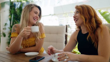 İki kadın evdeki terasta oturmuş kahve içiyor ve konuşuyor.