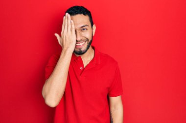 İspanyol sakallı, günlük kırmızı tişörtlü, bir gözünü eliyle kapatan, yüzünde kendine güvenen bir gülümseme ve sürpriz duygular olan bir adam.. 