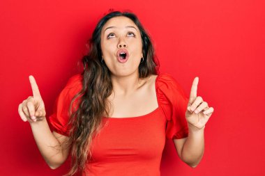 Gündelik kıyafetler giyen genç İspanyol kız şaşkın şaşkın yukarı bakıyor ve parmaklarıyla işaret ediyor ve kollarını kaldırıyor.. 