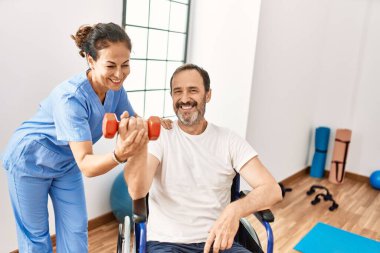 Orta yaşlı bir adam ve kadın, fizyoterapi kliniğinde tekerlekli sandalyede oturan dambılı kullanarak rehabilitasyon seansı yapıyorlar.