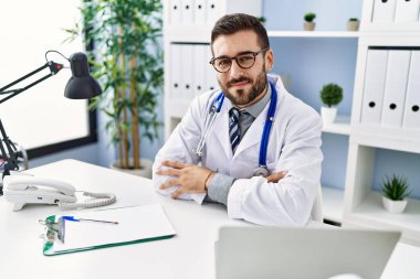 Young hispanic man wearing doctor uniform using laptop sitting at clinic