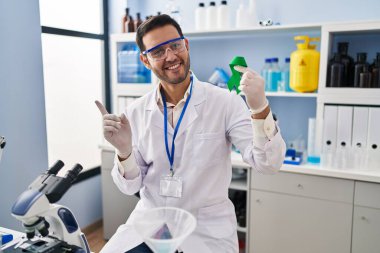 Sakallı, genç, labaratuarda çalışan, elinde yeşil kurdeleyle gülümseyen, eli ve parmağı kenarda gezen İspanyol bir adam. 