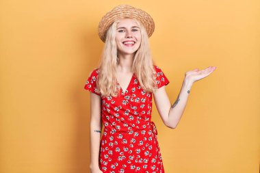 Beautiful caucasian woman with blond hair wearing summer hat smiling cheerful presenting and pointing with palm of hand looking at the camera. 