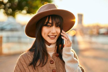 Kışlık şapkalı esmer kadın gün batımında açık havada telefonda konuşurken gülümsüyor.