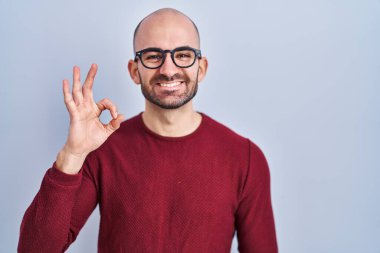 Beyaz arka plan üzerinde duran sakallı genç ve kel bir adam. Gözlüklü, gülümseyen bir adam. El ve parmaklarıyla 
