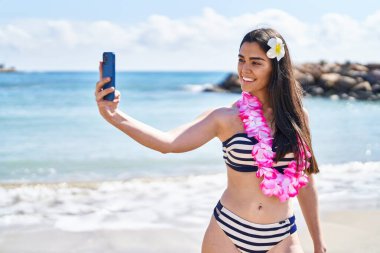 Bikini giyen genç İspanyol kadın deniz kenarında akıllı telefonla selfie çekiyor.