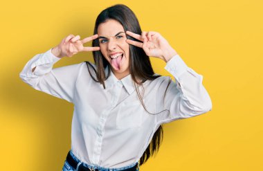 Young brunette teenager doing peace gesture close to eyes sticking tongue out happy with funny expression. 