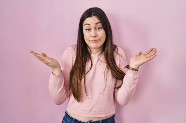 Pembe arka planda duran esmer bir kadın. Kolları ve elleri havada, kafası karışmış bir ifade. şüphe kavramı. 