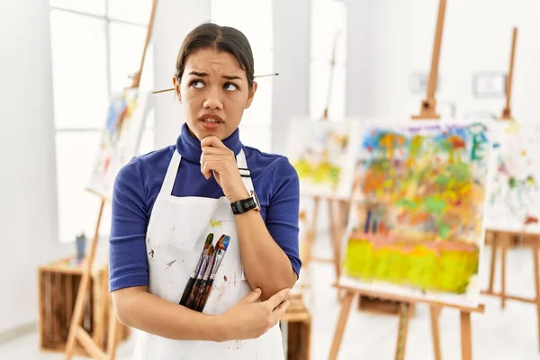 Young brunette woman at art studio thinking worried about a question, concerned and nervous with hand on chin