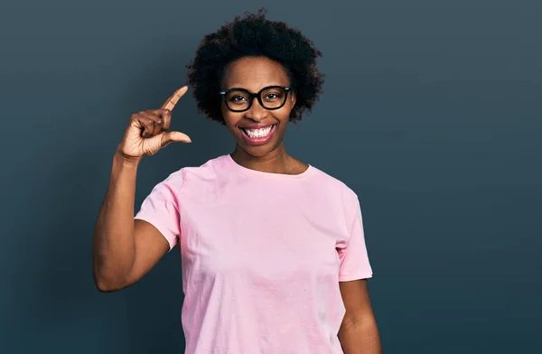 African american woman with afro hair wearing casual clothes and glasses smiling and confident gesturing with hand doing small size sign with fingers looking and the camera. measure concept.