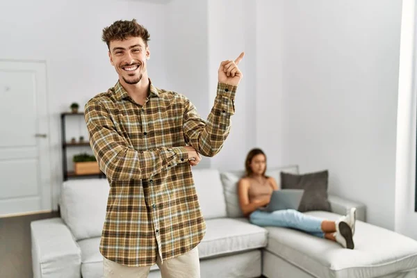 Young handsome man at the living room with girlfriend smiling happy pointing with hand and finger to the side
